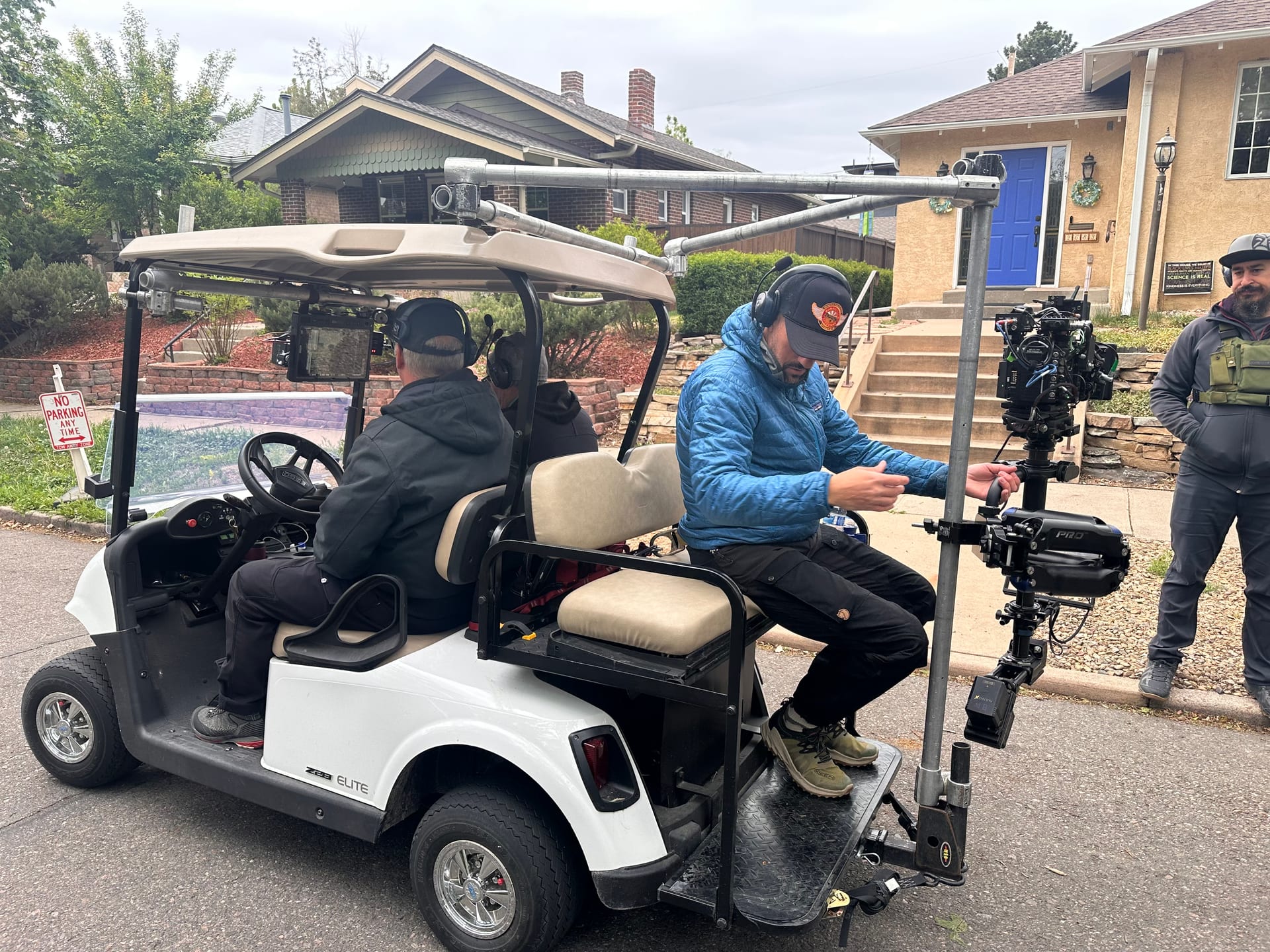 Interior crew positions on the grip cart camera car: driver and operator wearing headsets, with a rear-mounted stabilized cinema camera rig and an onboard monitor, parked on a suburban street between houses.